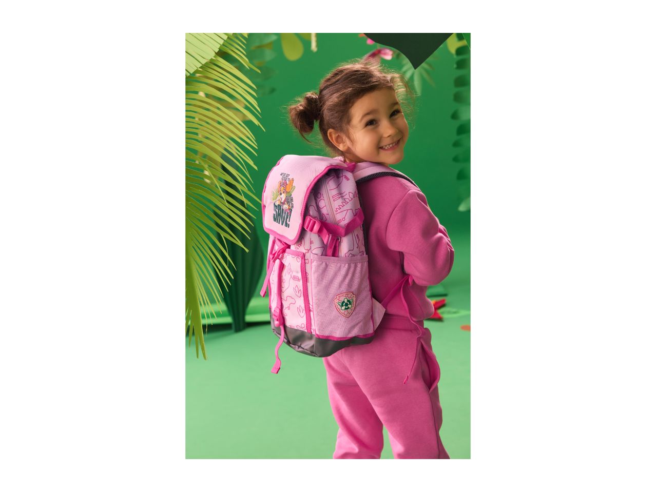 Smiling girl in pink tracksuit and backpack with cartoon dog in a jungle setting.