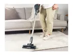 Person vacuuming a light-colored rug in a living room with a cordless stick vacuum.