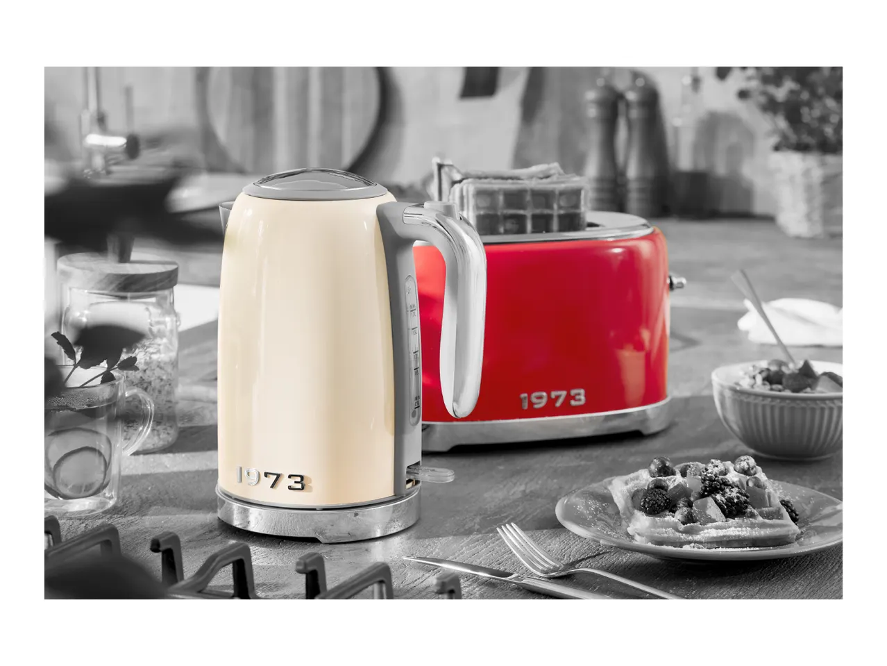 Cream kettle and red toaster with '1973' branding, waffles, and tea on a kitchen counter.