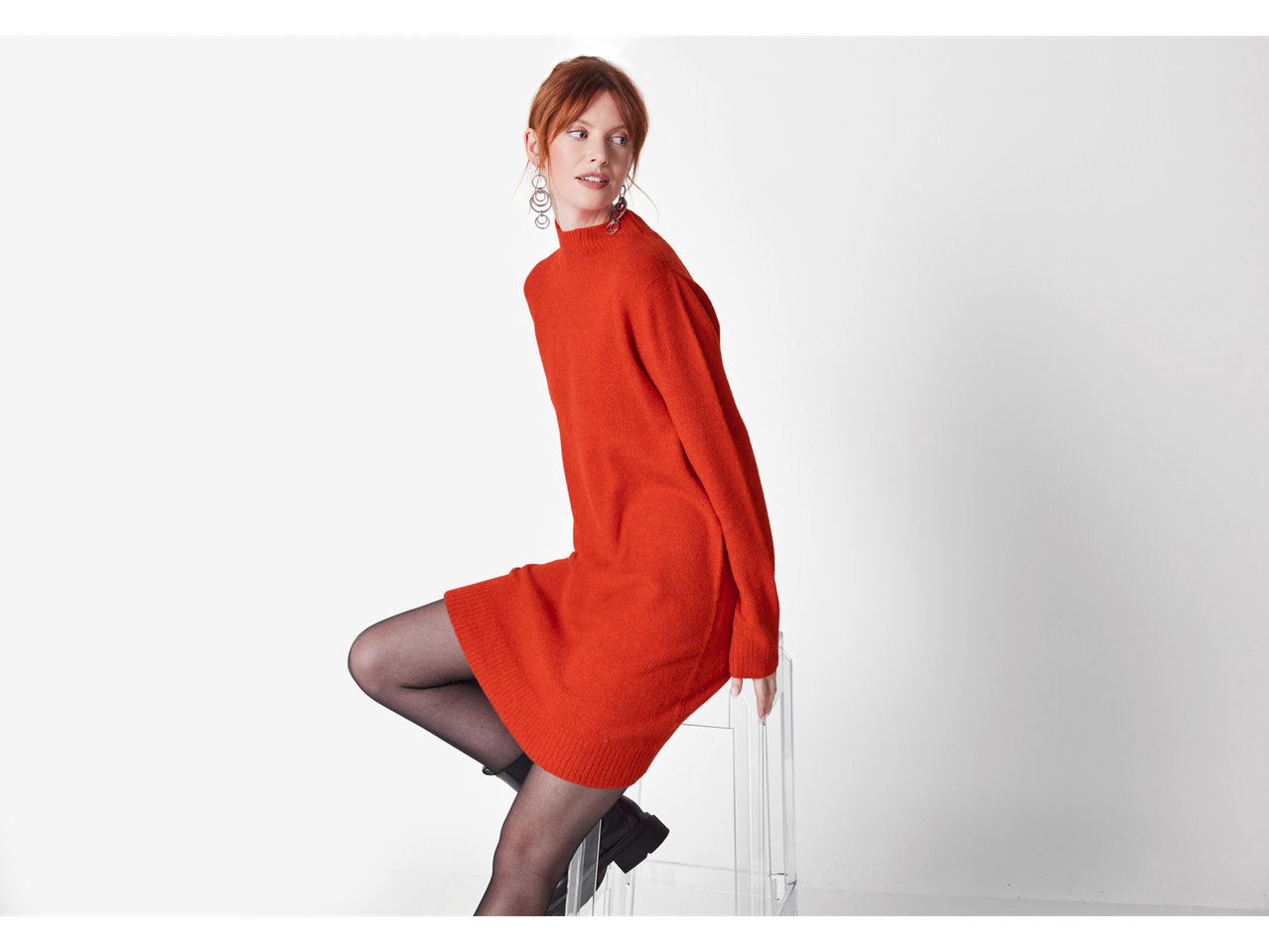 A woman in an orange Esmara ladies' jumper dress and black tights sits on a clear stool.