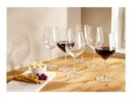 Six wine glasses, some filled with red wine, on a wooden surface with cheese, grapes, and breadsticks.