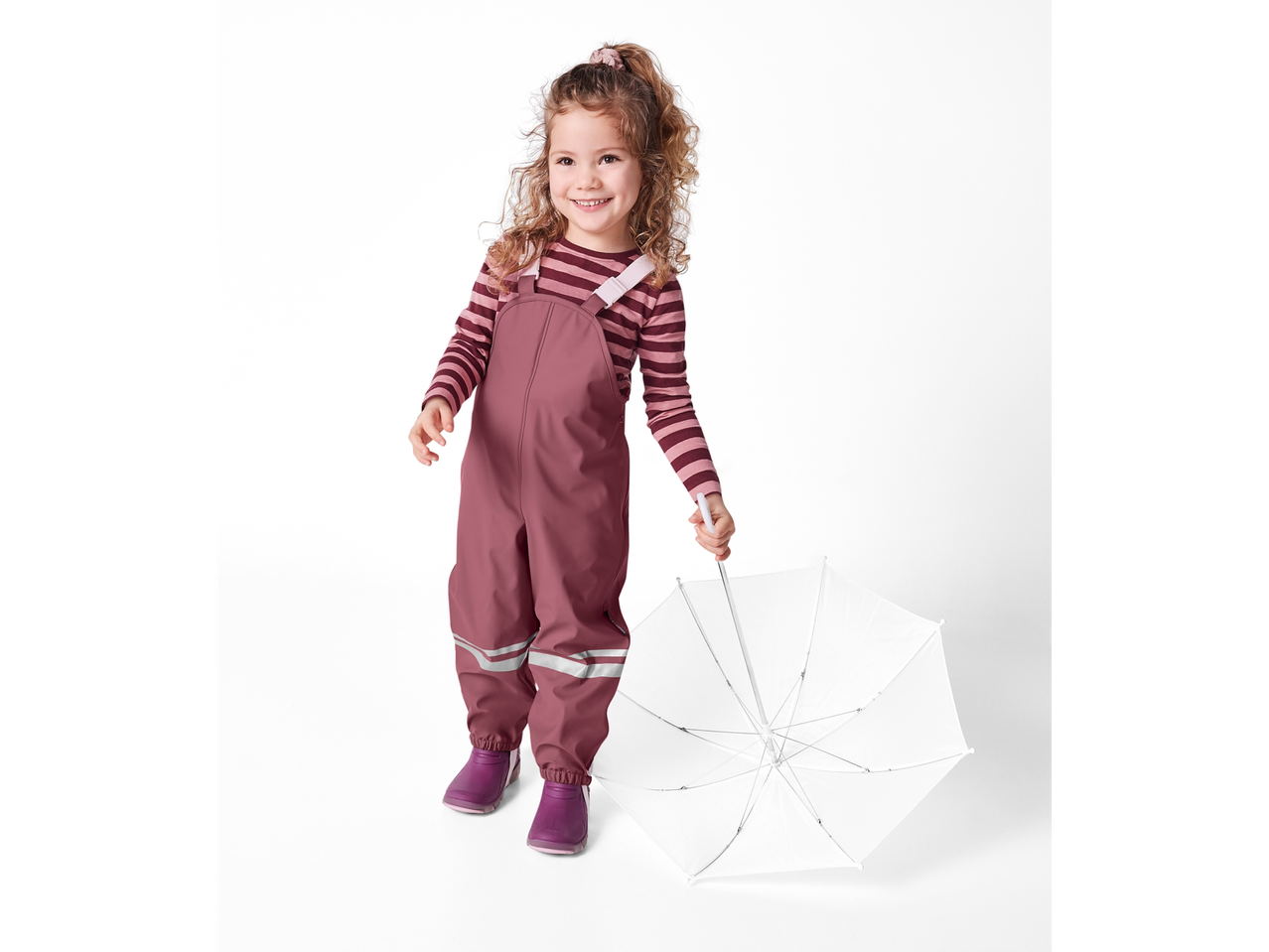 O fetiță zâmbitoare în pantaloni și cizme de ploaie Reima ține o umbrelă albă.