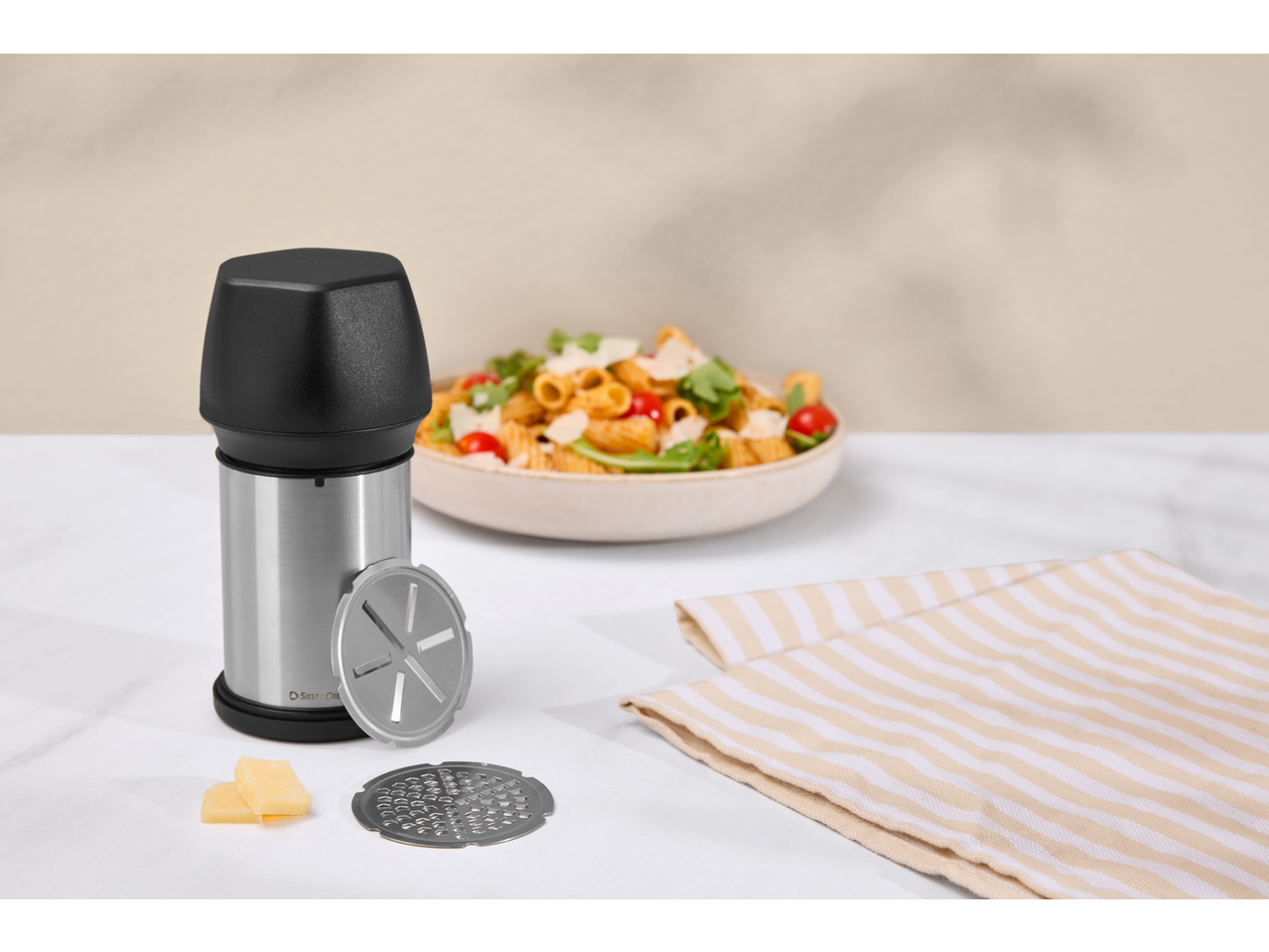 Răzătoare de brânză cu paste, brânză și un prosop dungat pe o masă albă.