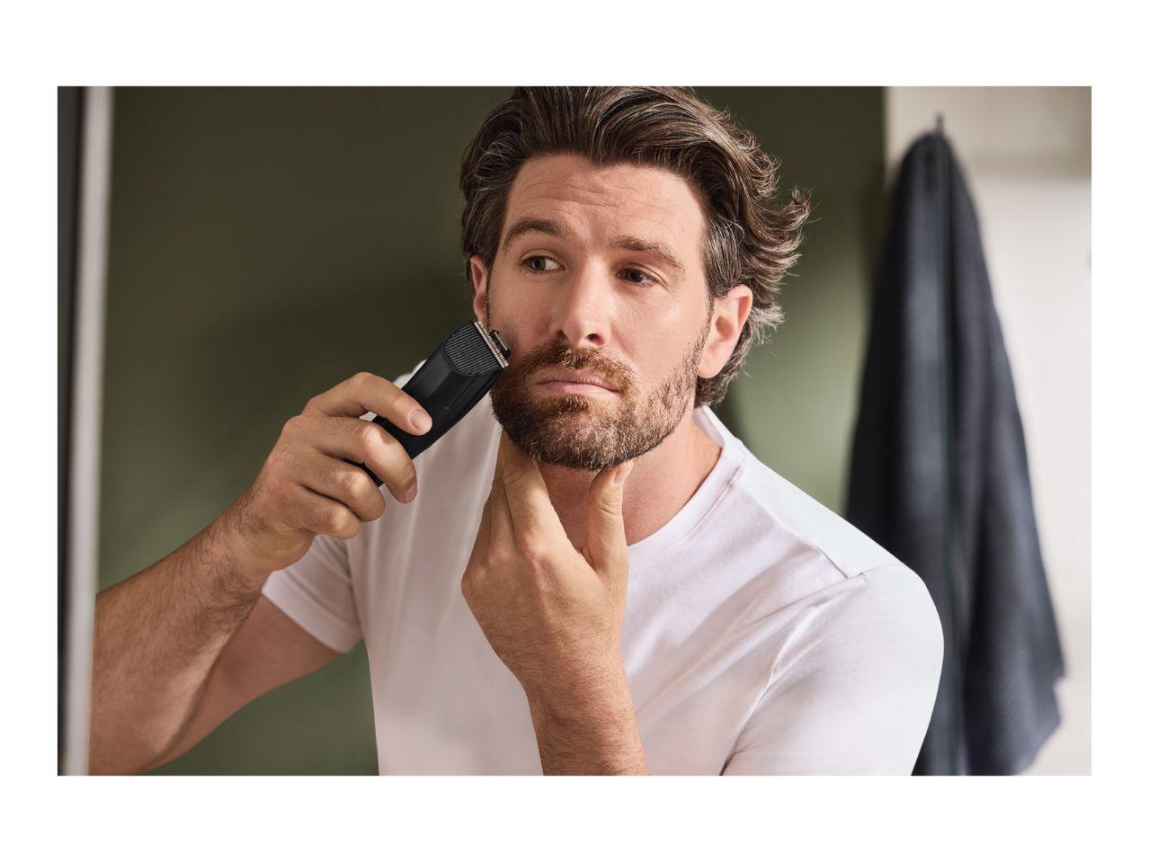 Man trimming his beard with a beard trimmer in a bathroom mirror.