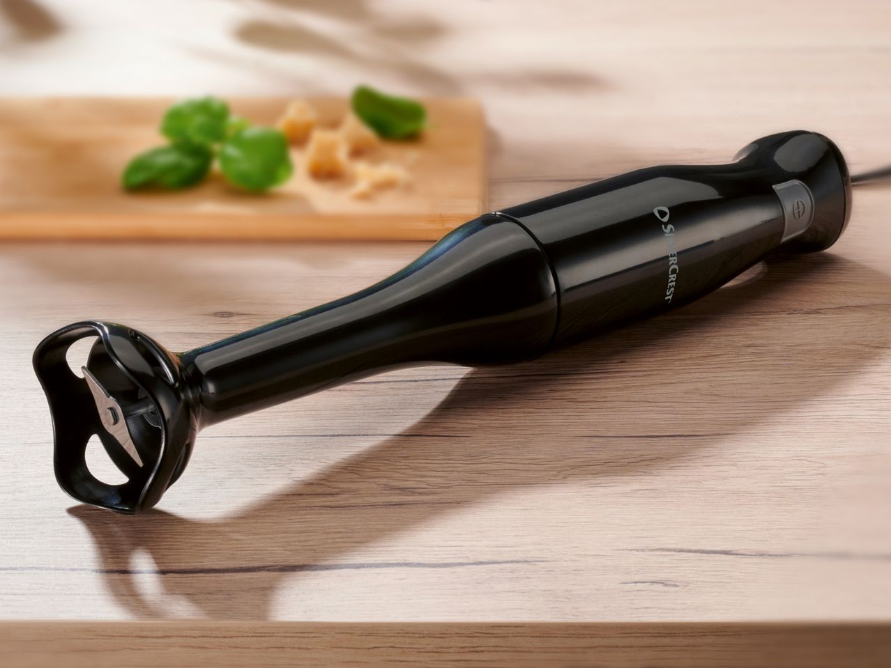 Black Silvercrest immersion blender on a wooden surface with basil and cheese in the background.