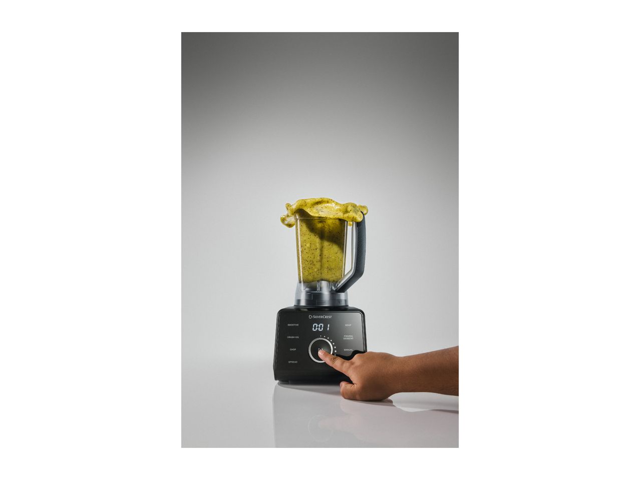 A hand pressing the start button on a black blender filled with a green smoothie.