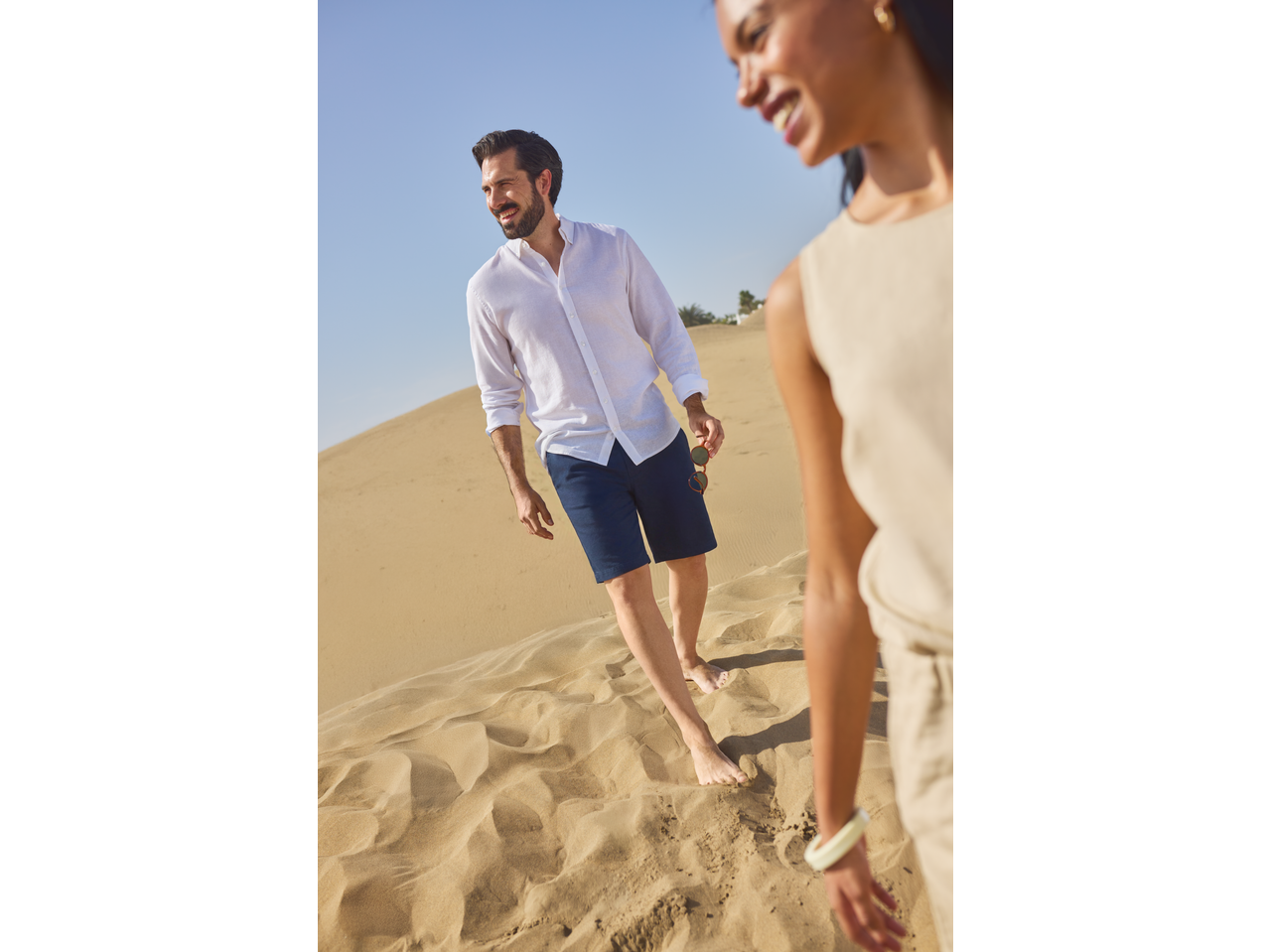 Man in white linen shirt and navy shorts walking barefoot on sand dunes with a woman.