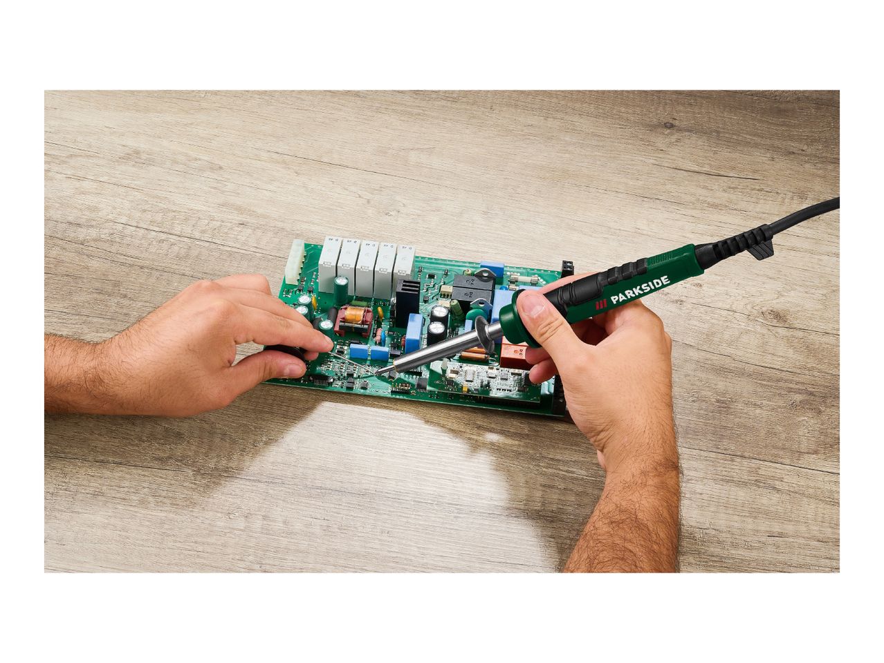 Hands soldering a circuit board with a soldering iron on a wooden surface.