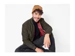 Man in a dark green bomber jacket, black turtleneck, and brown beanie smiling.