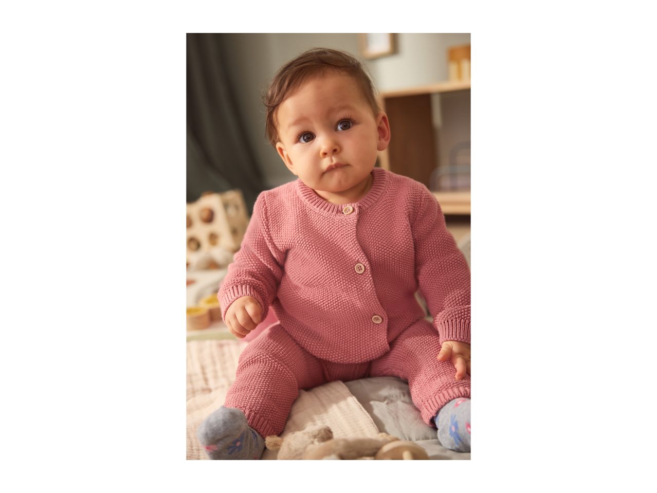 Baby wearing a pink knitted cardigan and trousers, sitting on a blanket.