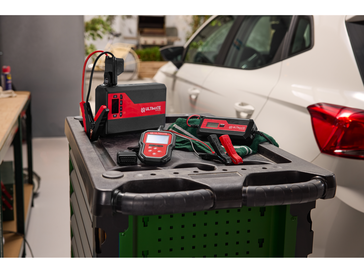 ULTIMATE SPEED® OBD 2 diagnostic devices and battery charger on a trolley next to a white car.