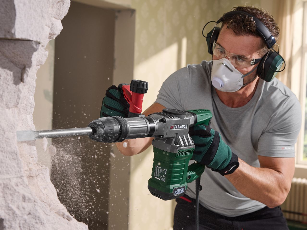 Man in safety gear using a demolition hammer to break a wall, creating dust.