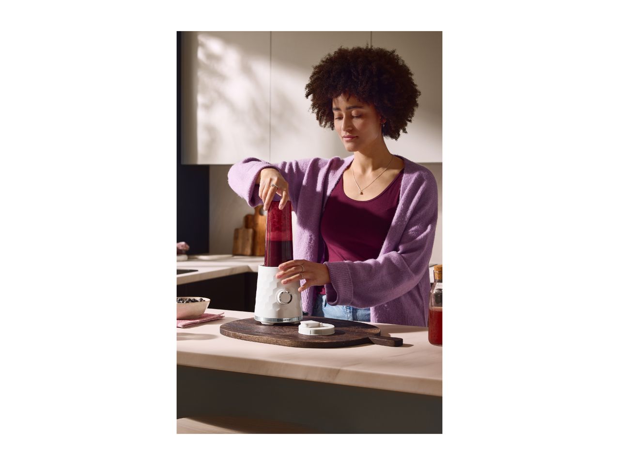 A woman using a compact white blender to make a berry smoothie in a kitchen.