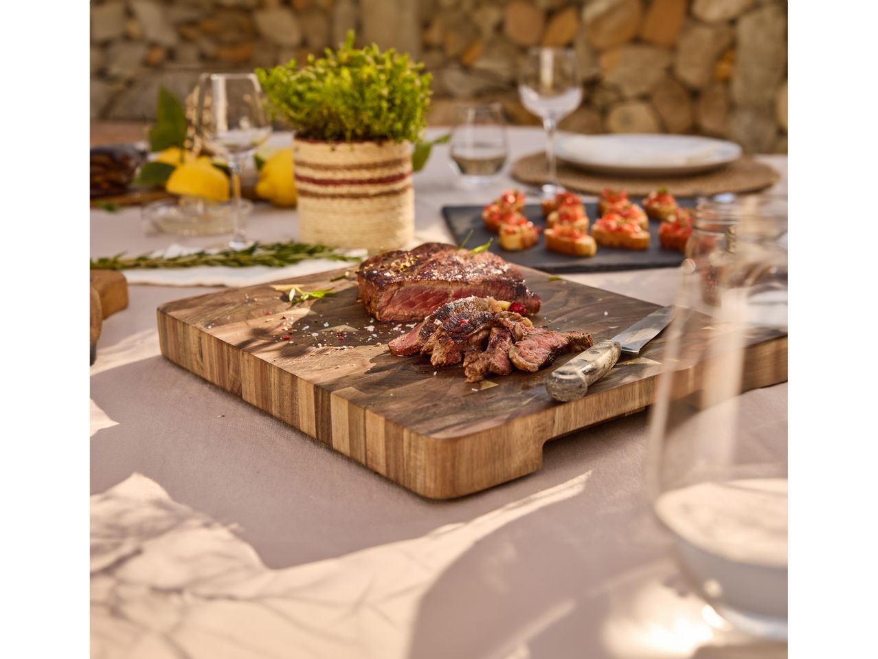 A rustic wooden cutting board displays a roasted meat, set on a table with other food and drink.