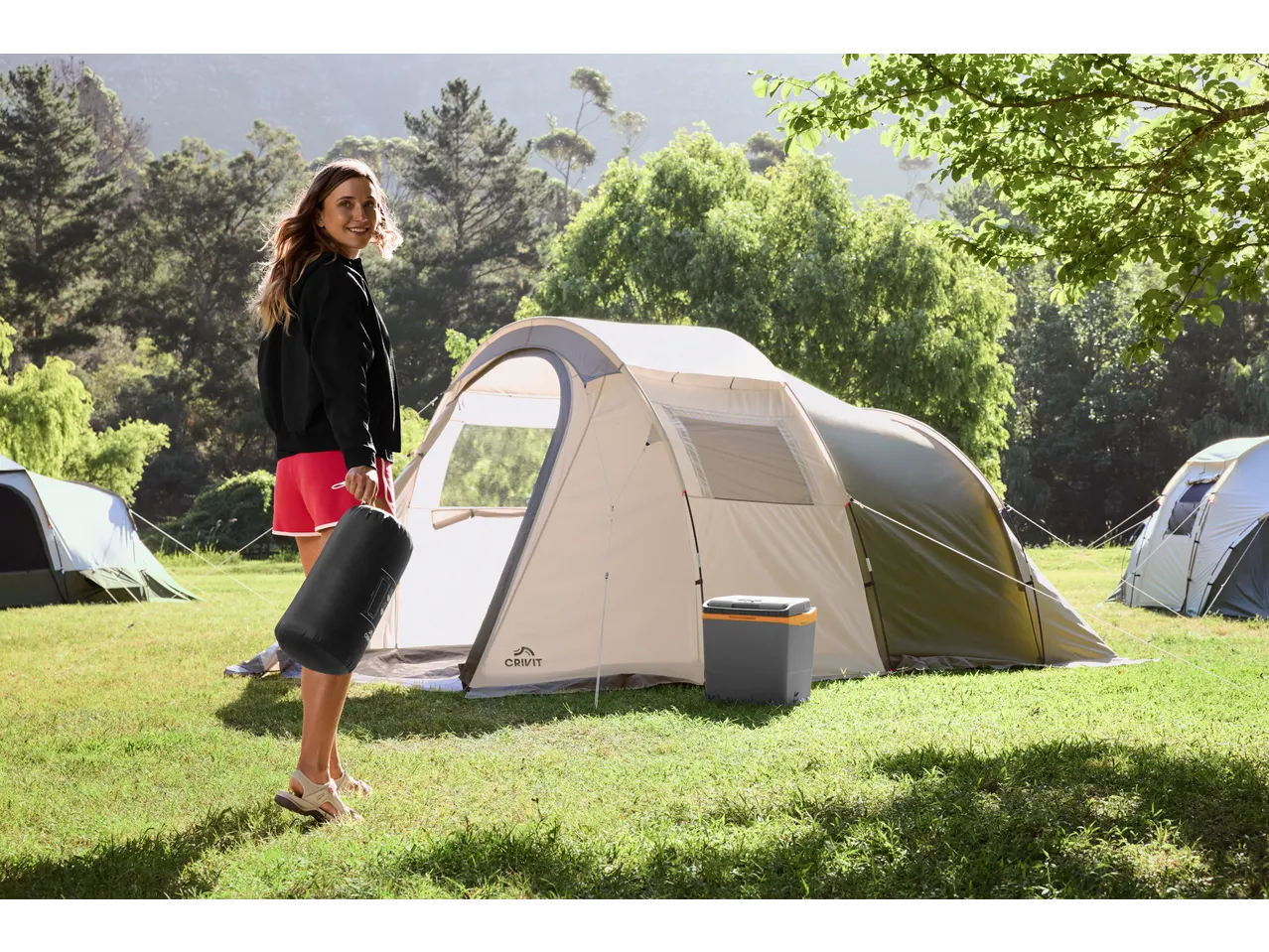 Woman holding a sleeping bag next to a large camping tent and a cooler in a sunny field.
