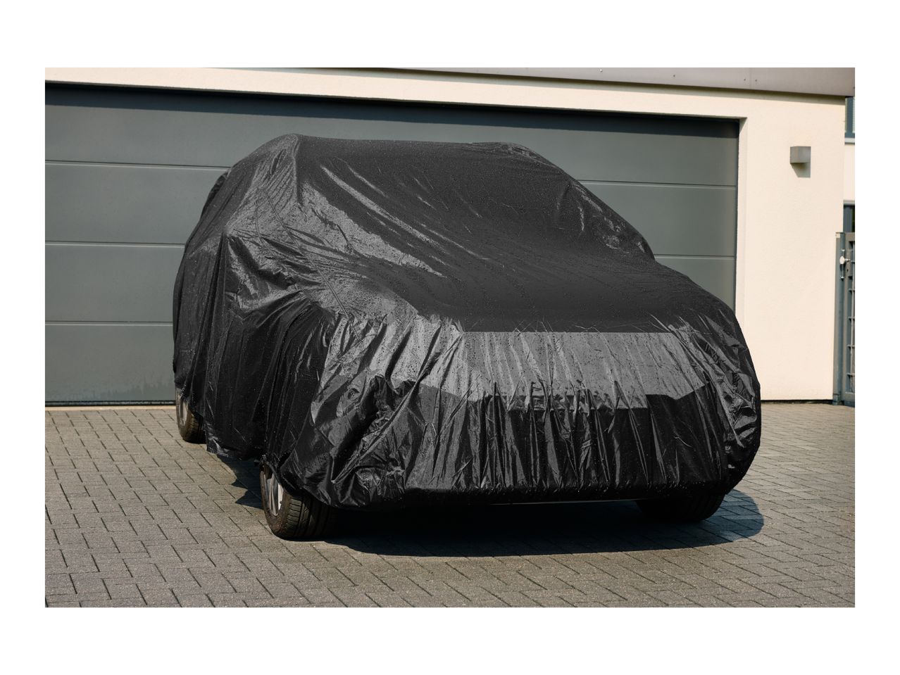 Black car cover on a car parked on a paved driveway in front of a garage.