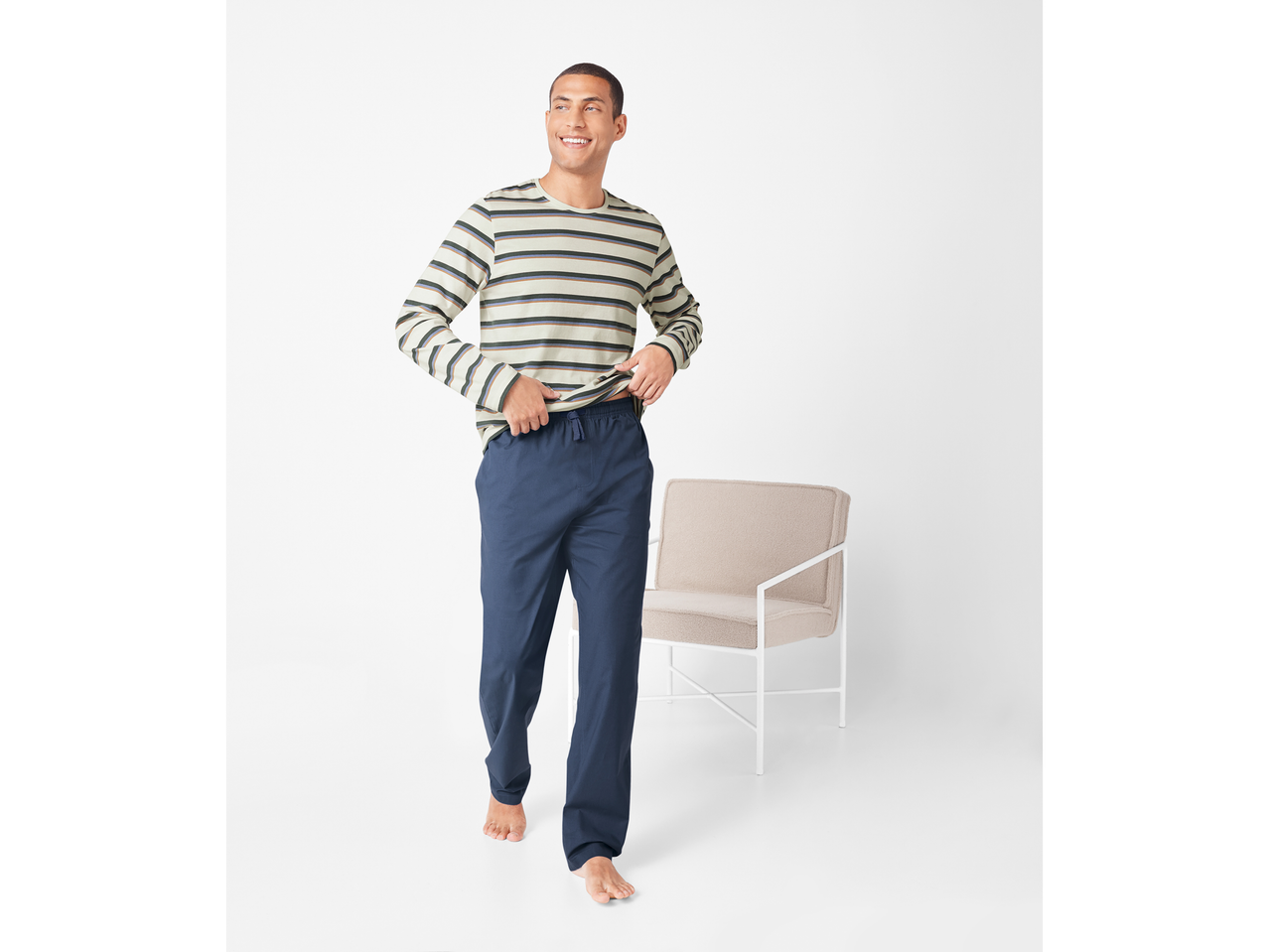 Bărbat zâmbitor în pijama cu dungi și pantaloni albaștri, lângă un scaun bej.