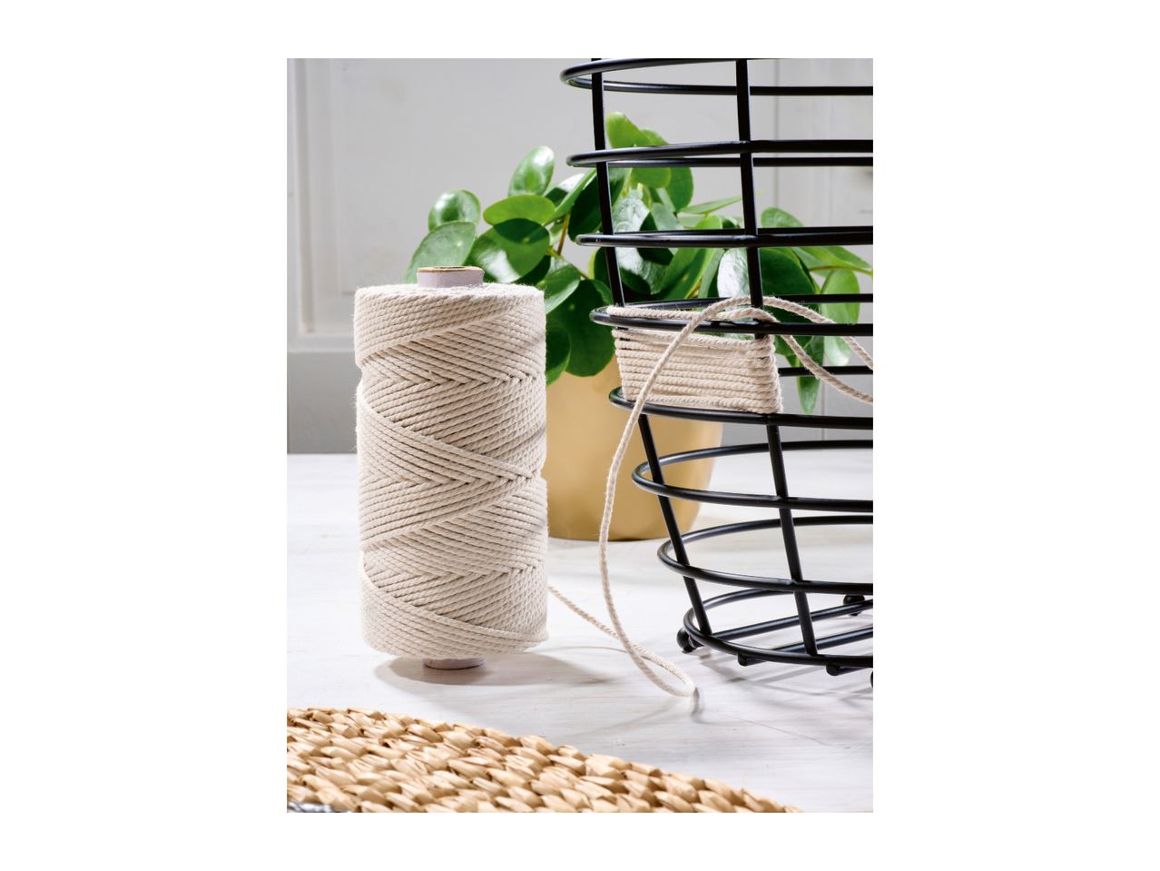 A spool of macrame cord next to a black wire basket being wrapped with the string.