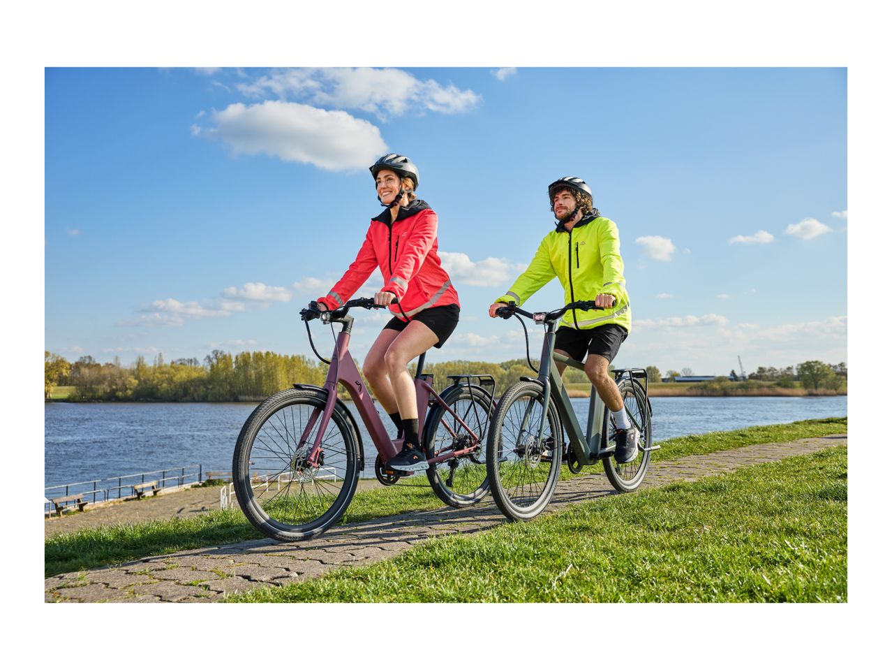 Two people cycling on electric bikes along a river path on a sunny day.