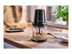 A food chopper with cashews on a wooden counter, with a bowl of cashews and a plant in the background.