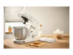 Cream stand mixer with whisk attachment, bowl, and baking ingredients on a wooden counter.