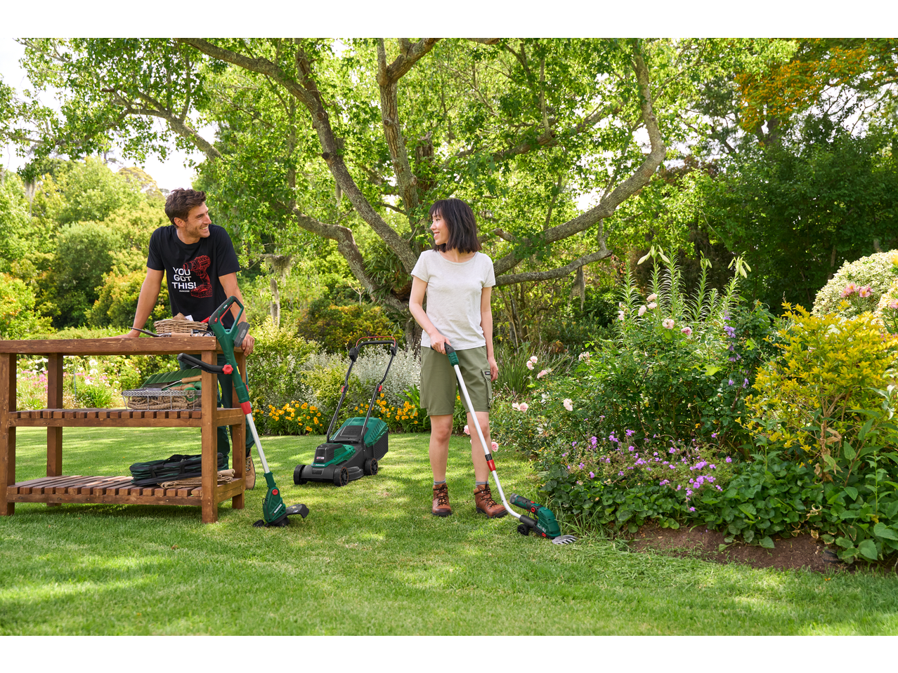 Man and woman in a garden with a lawnmower, grass trimmer, and hedge trimmer.