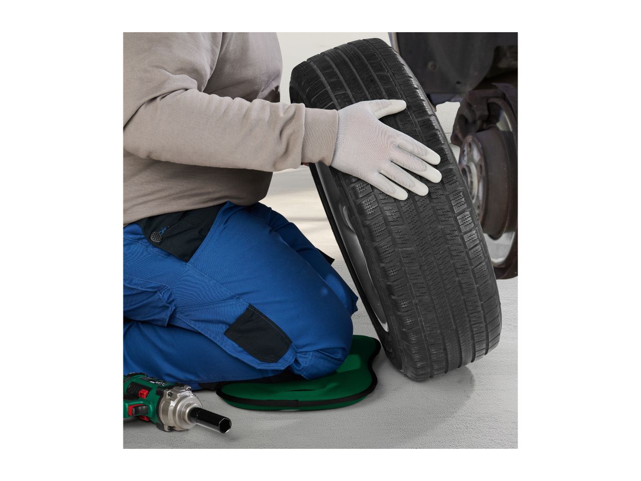 A mechanic kneeling on a green pad while changing a car tire with an impact wrench nearby.