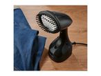 A black handheld garment steamer with a brush attachment next to blue jeans on a wooden surface.
