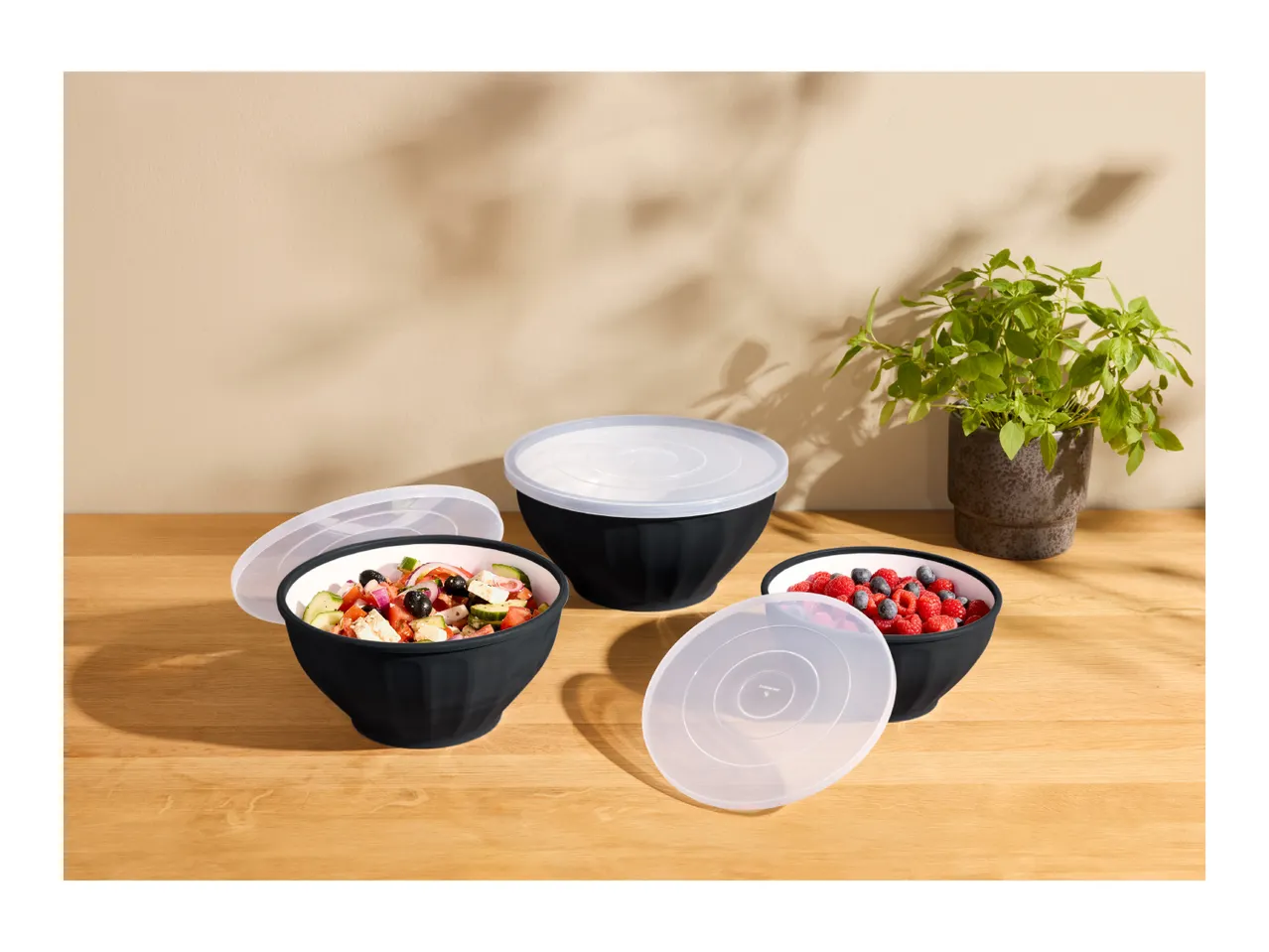 Black bowls with lids, one with salad, one with berries, on a wooden table.