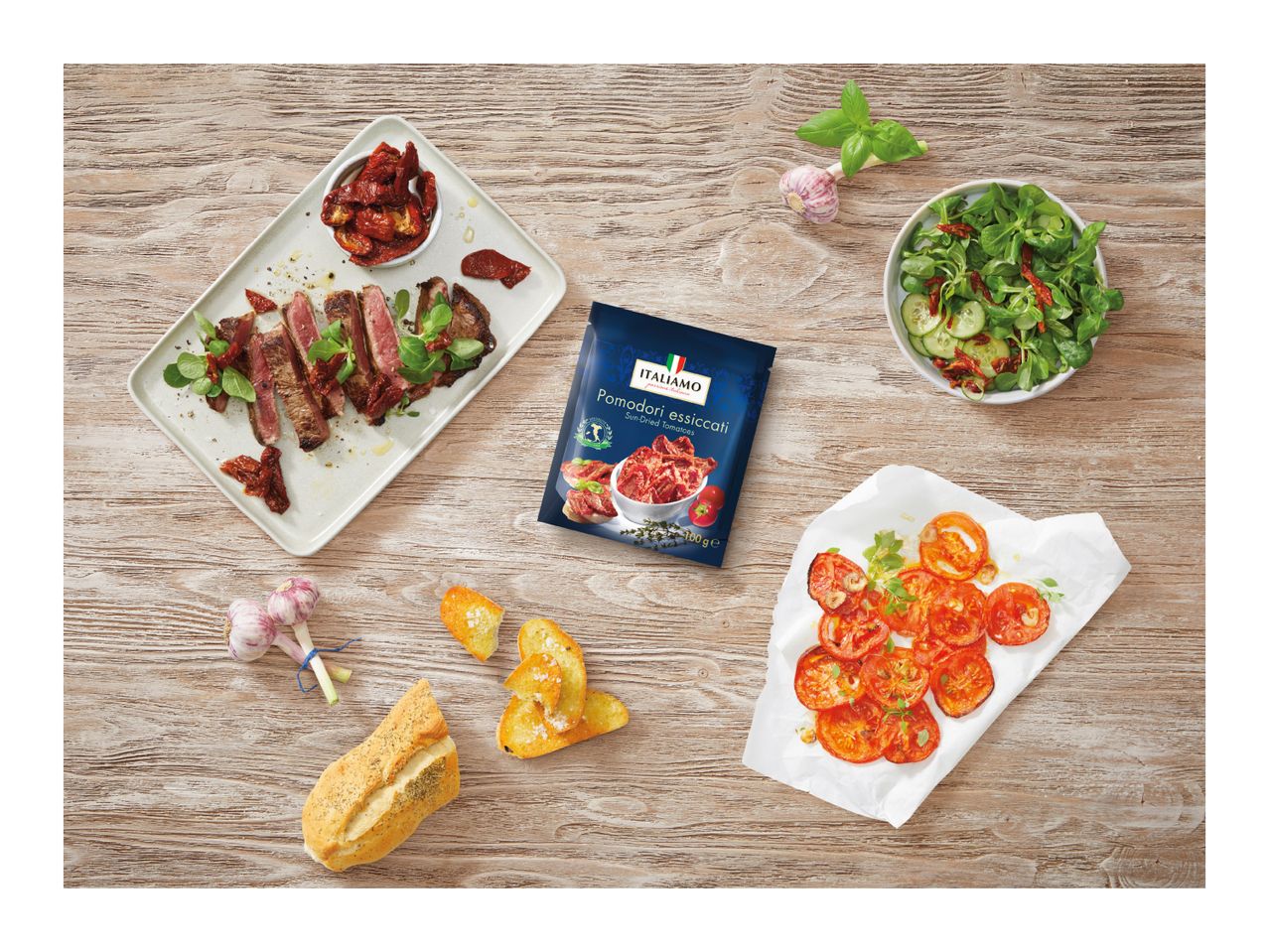 A blue packet of sun-dried tomatoes surrounded by steak, salad, bread, and garlic on a wooden table.