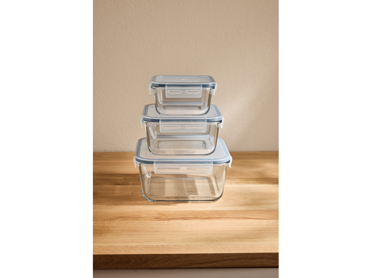 Stack of three rectangular glass food storage containers with light blue lids on a wooden surface.
