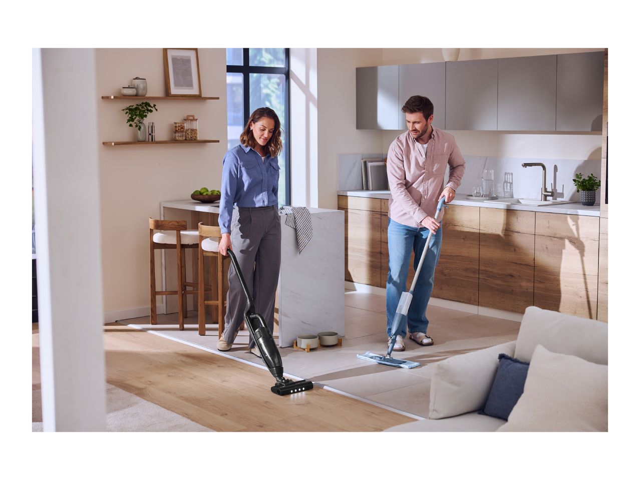 A woman vacuums a wooden floor while a man mops a tiled floor in a modern kitchen.