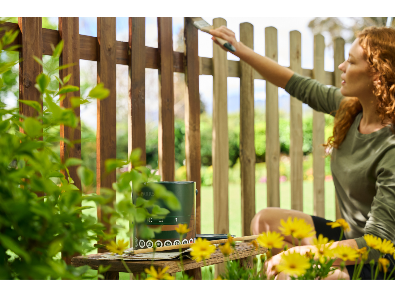 Woman painting a wooden fence with a Parkside® paintbrush, paint can on a stool.