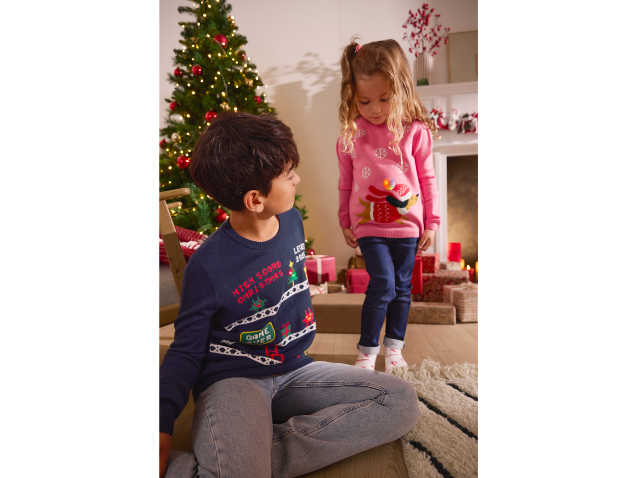 Two children in Lupilu light-up Christmas jumpers, boy in blue, girl in pink, by a Christmas tree.