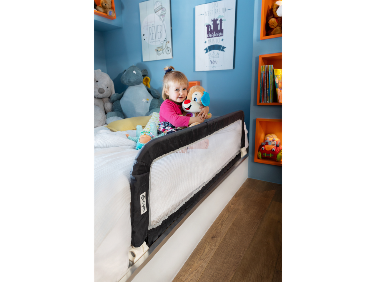 A smiling toddler with a plush toy in a bed with a Safety 1st bed rail.
