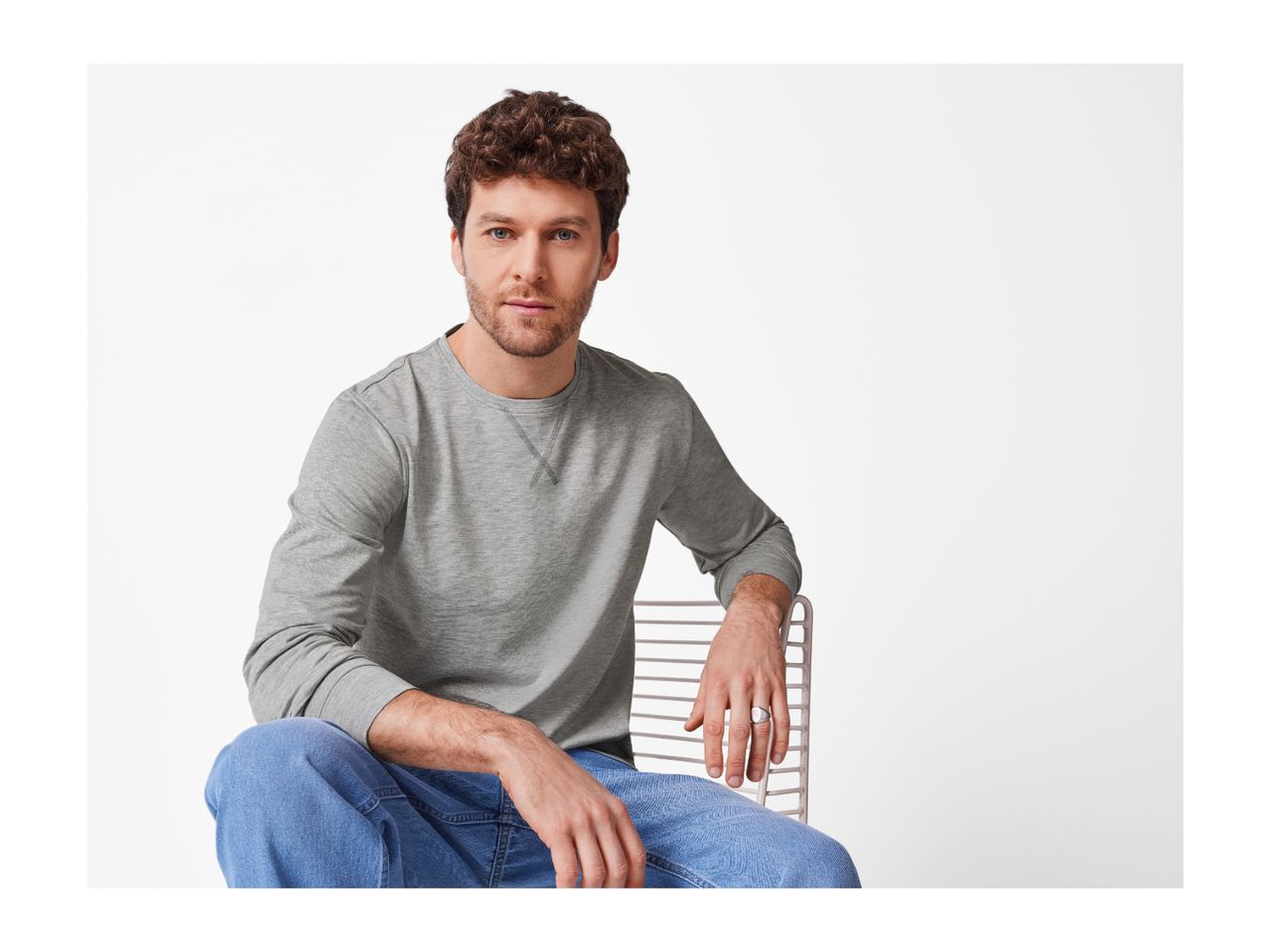 A man in a gray long-sleeve shirt and blue jeans sits on a chair.