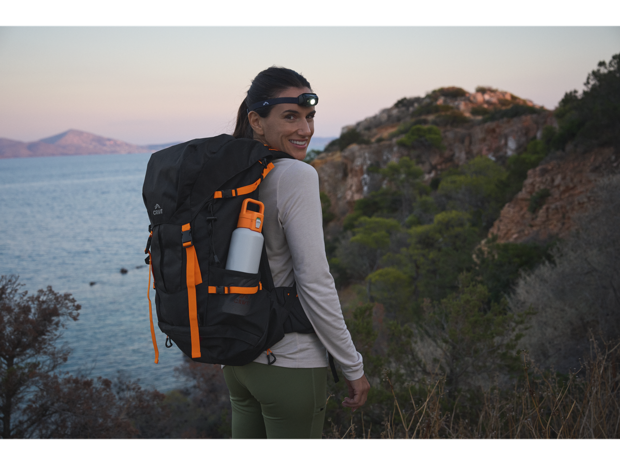 Femeie cu rucsac Crivit și lanternă frontală, zâmbind pe fundalul mării și munților.
