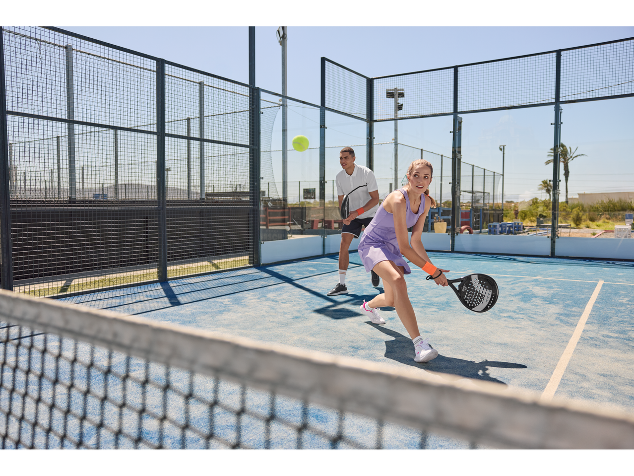 Jogadores de padel numa quadra azul com raquetes e bola.