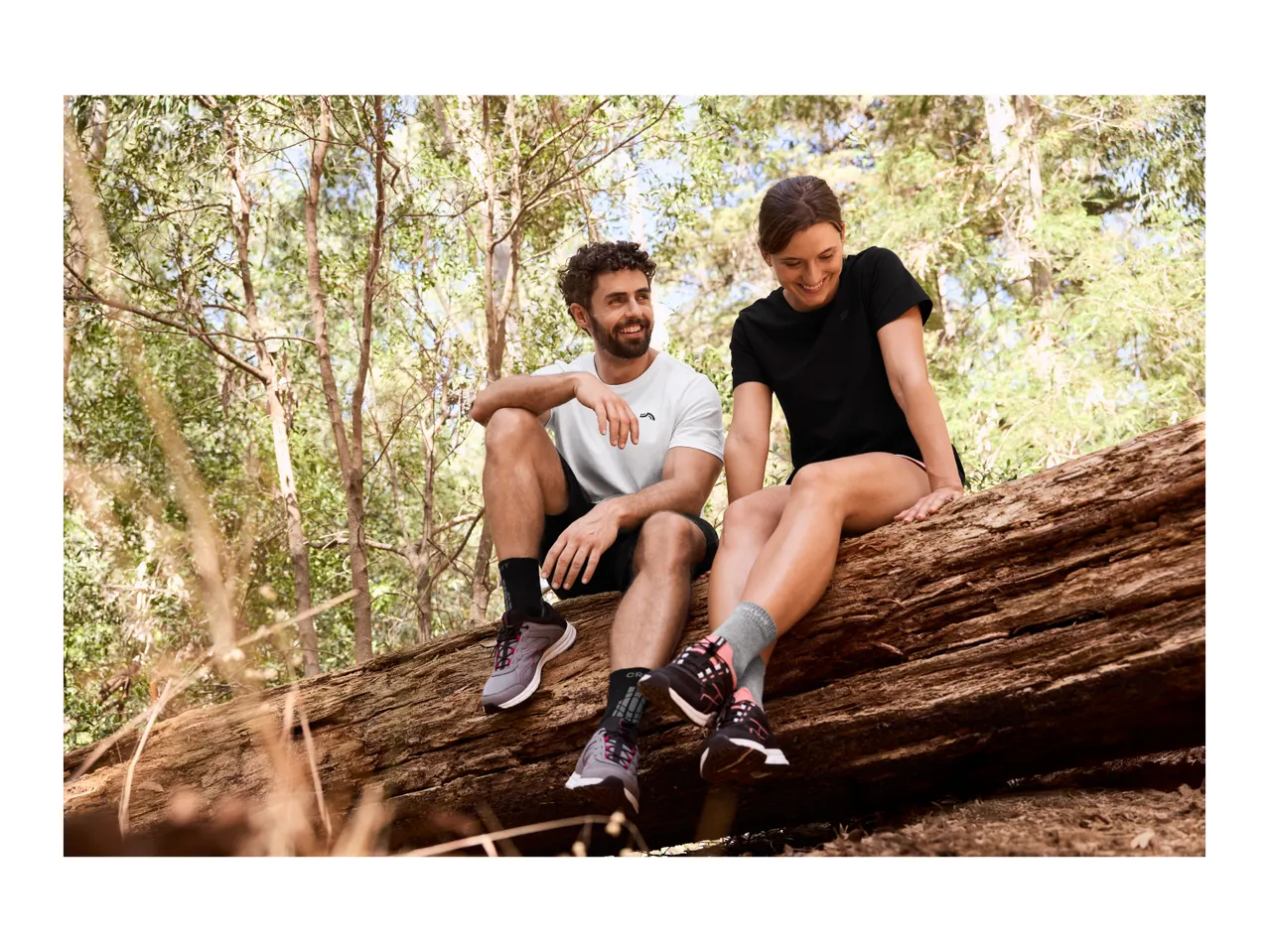 Two people in activewear sitting on a fallen log in a forest, smiling.