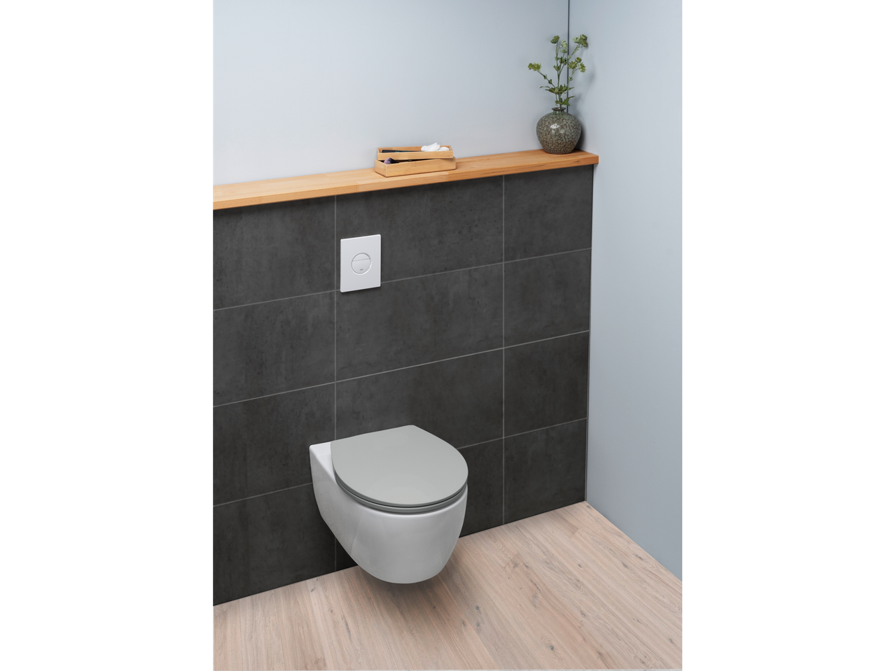 A modern toilet with a gray seat, flush plate, and a wooden shelf with decor.