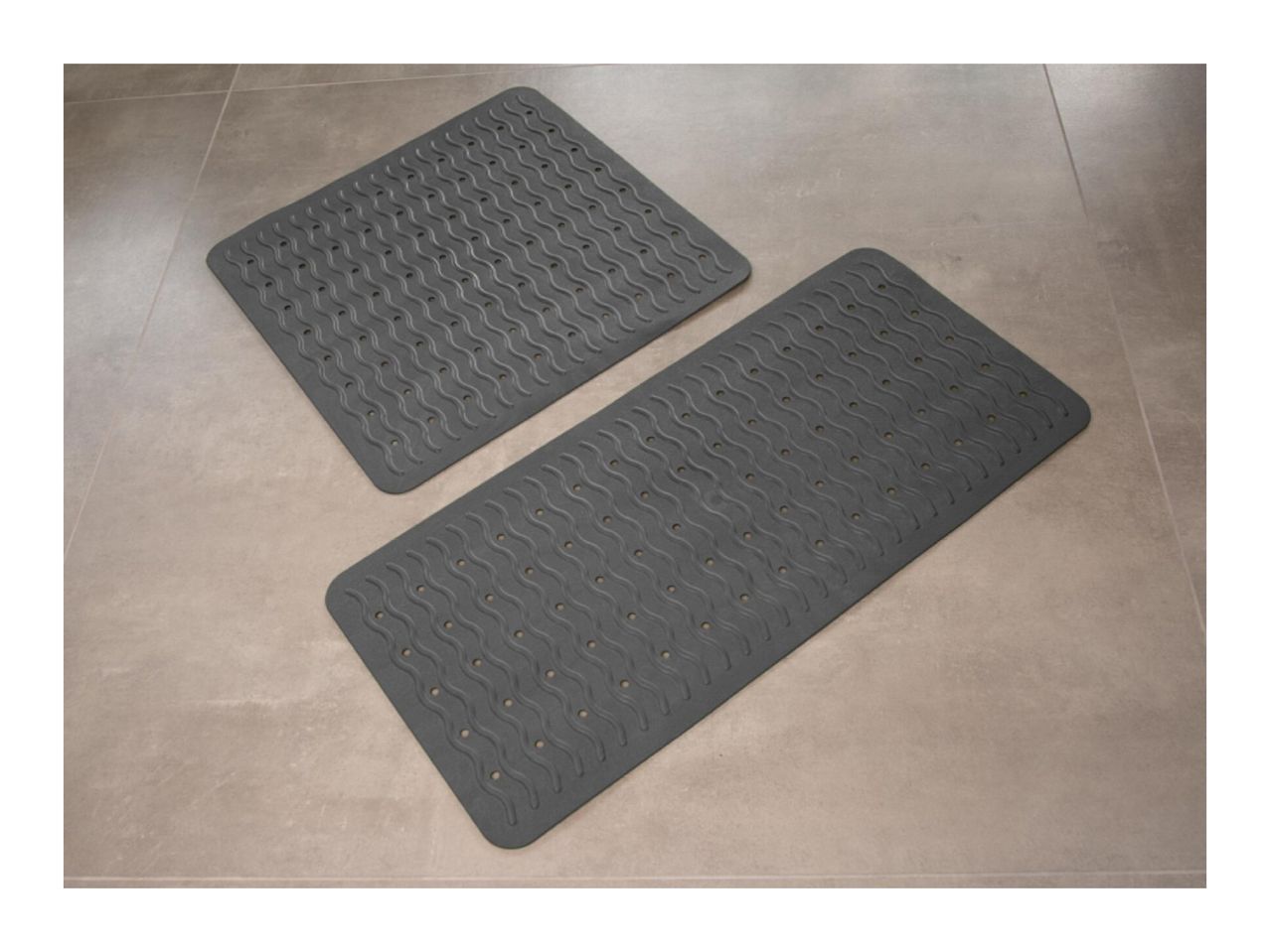 Two dark gray bath mats with wavy patterns and drainage holes on a tiled floor.