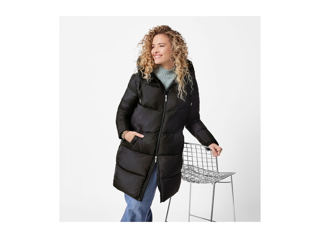 Woman in a black long puffer jacket, light blue sweater, and jeans, smiling and holding a chair.