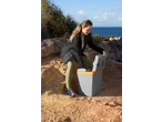 Woman opening a portable cooler on a rocky path by the sea.