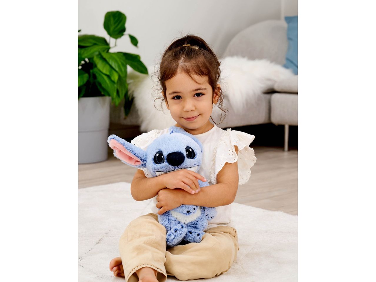 A young girl sitting on a rug, holding a blue Stitch plush toy.