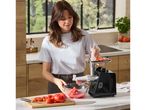 Woman using a black electric meat grinder to process tomatoes into a clear bowl on a kitchen counter.