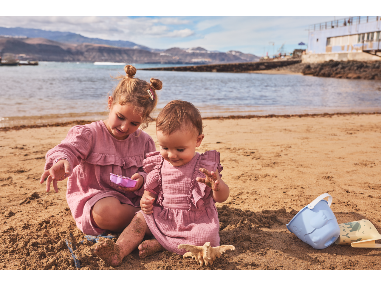 Două fete în rochii de muselină LUPILU jucându-se în nisipul plajei.