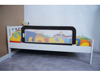 White toddler bed with a black bed rail, colorful bedding, and a teddy bear.