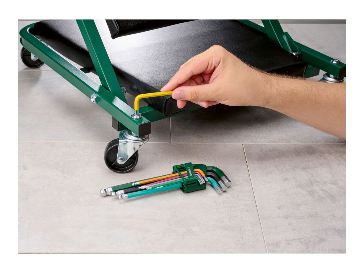Person assembling a green workshop creeper with a yellow hex key, with a set of hex keys nearby.
