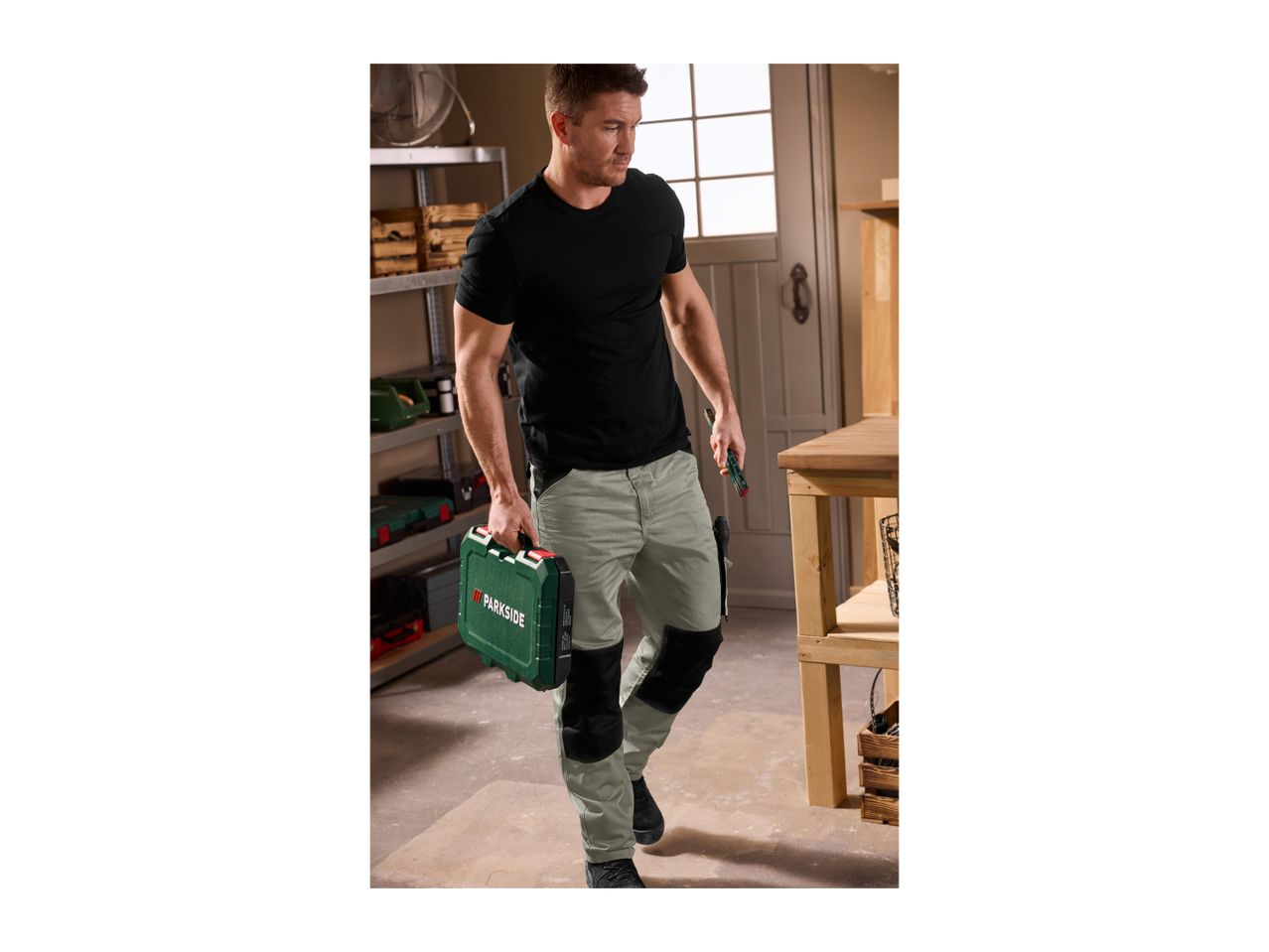 Man in work trousers carrying a toolbox and a utility knife in a workshop.