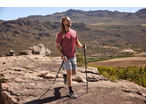 Man in a red t-shirt and grey shorts hiking with poles on a rocky mountain path.